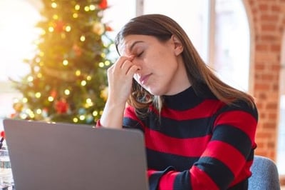 woman sitting at the table working with laptop at home around christmas tree tired rubbing nose and eyes feeling fatigue and headache. Stress and frustration concept.