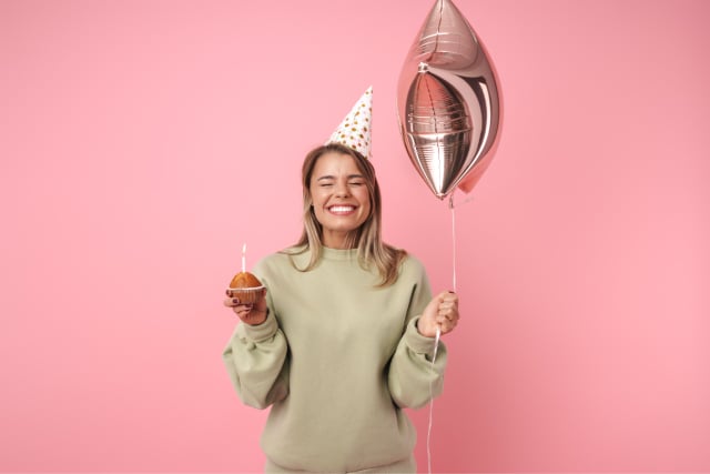 Excited person wearing birthday hat holding balloon and cupcake