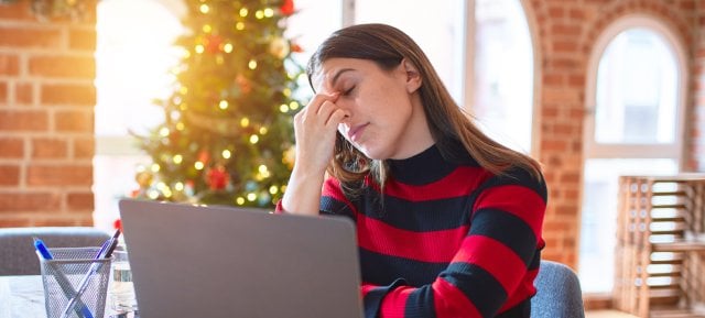 woman sitting at the table working with laptop at home around christmas tree tired rubbing nose and eyes feeling fatigue and headache. Stress and frustration concept.